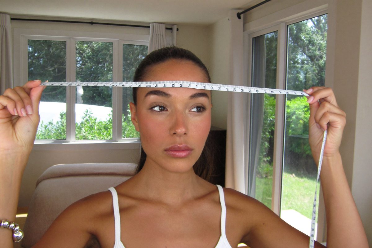 Woman measuring her face shape at home with a soft tape measure across her forehead, hair pulled back